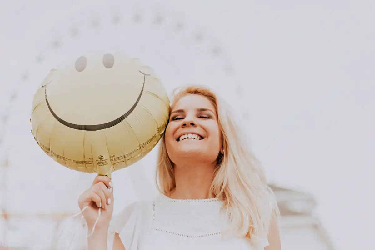 Woman smiling holding a smiley face balloon - Happy Mind Training Blog | New Month Resolutions