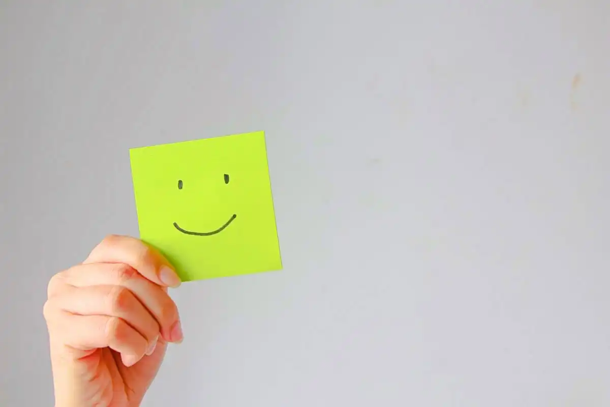 Hand holding a sticky note with a smiley face drawn on it - Happy Mind Training Blog | Compliments Make Us Feel Good